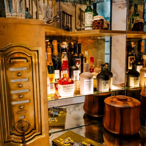 A cozy bar display with liquor bottles, a Maker&rsquo;s Mark whisky, and cocktail tools behind glass and copper tubing.