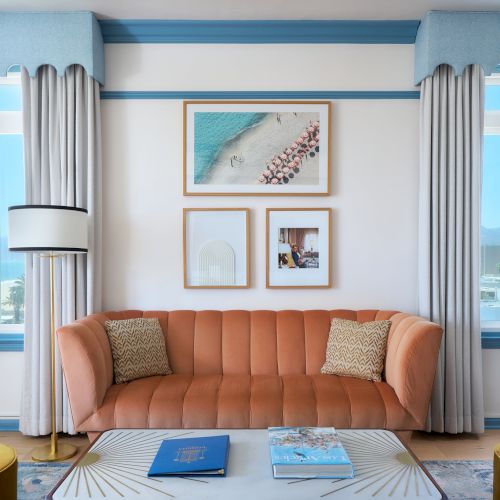 A bright living room with an orange sofa, gray curtains, framed artwork, and large windows showing a distant sea view.