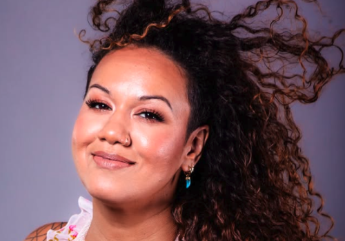 A smiling woman with long curly dark hair, wearing a floral pink top and small earrings against a soft studio backdrop.