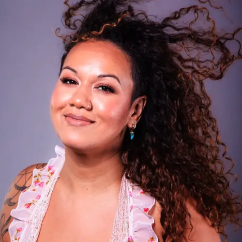 A smiling woman with long curly dark hair, wearing a floral pink top and small earrings against a soft studio backdrop.