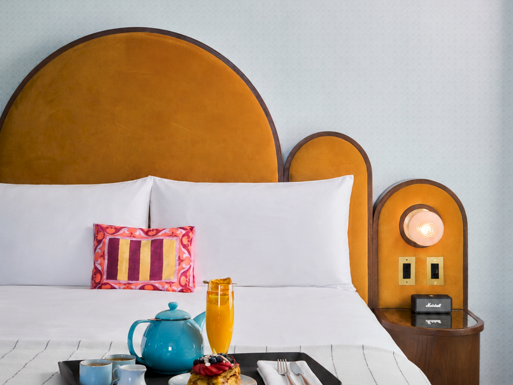 A cozy hotel room scene: a neatly made bed with pillows, a breakfast tray with tea and pastries, and a bedside lamp.