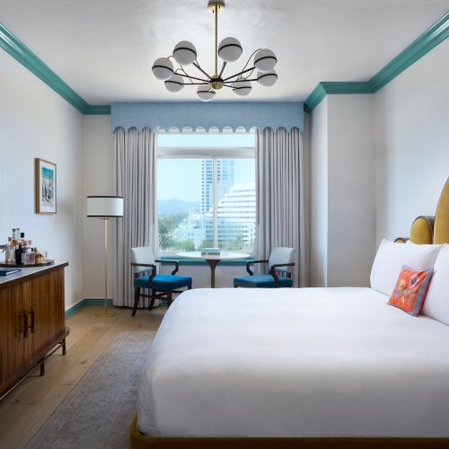 A bright hotel room with a king bed, chandelier, TV and desk, plus a window view over buildings and mountains.