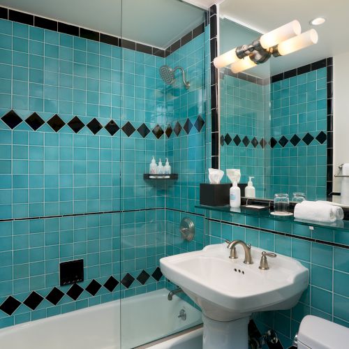 A teal-tiled bathroom with a glass shower, rain showerhead, vanity sink, towels, and soap bottles.