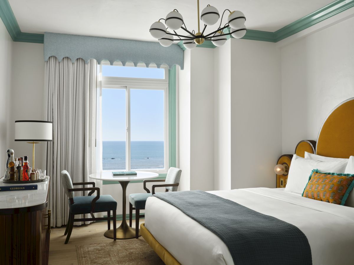 Bright seaside hotel room with a chandelier, ocean-view window, curtains, small table, and a neatly made bed.