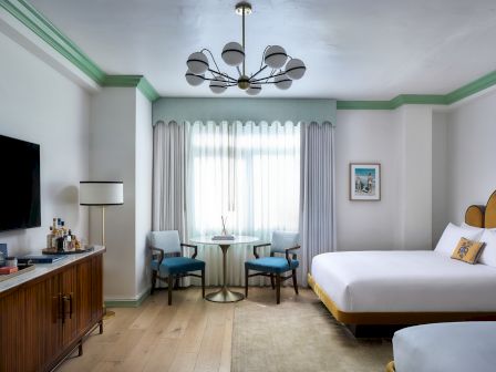 A bright hotel-style room with a chandelier overhead, a round table with chairs, curtains, and a TV and dresser on the left.
