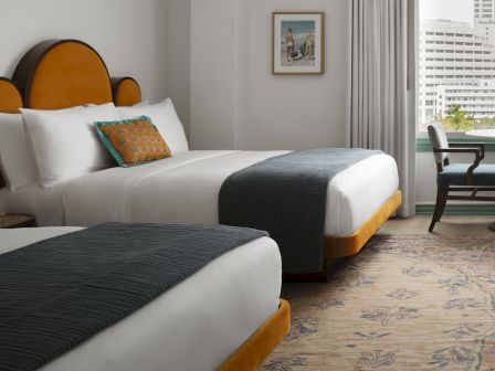 A bright hotel room with twin beds, colorful pillows, a framed beach photo, and a window overlooking city buildings.