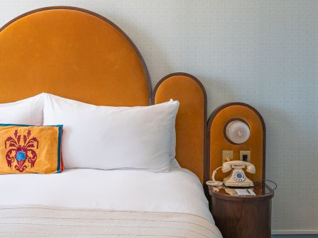 A hotel room bed with white pillows, a wooden arched headboard, and a vintage rotary phone on a side table.