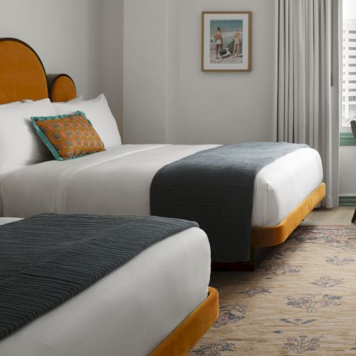 A bright hotel bedroom with a large bed, patterned pillows, a chair by the window, city skyline view, and framed beach art.