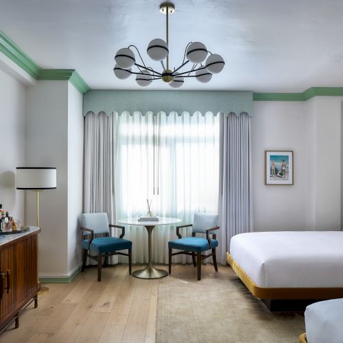 Bright modern room with a chandelier, round table and chairs, tall curtains, a TV console, and a bed with pillows.