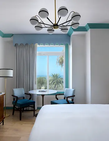 Bright hotel room with teal chandelier, large window and curtains, small table/chairs, TV desk, and a neatly made bed.