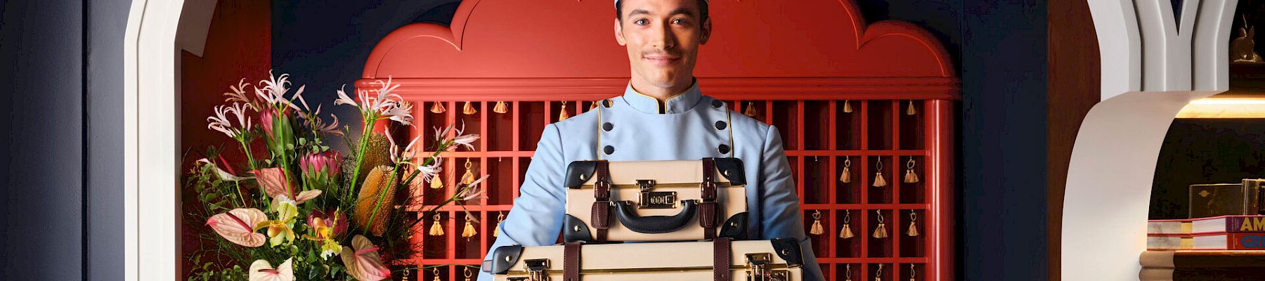A waiter in a light blue uniform carries stacked vintage suitcases in a stylish lobby with flowers and a curved red backdrop.