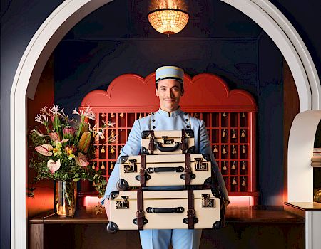 A waiter in a light blue uniform carries stacked vintage suitcases in a stylish lobby with flowers and a curved red backdrop.