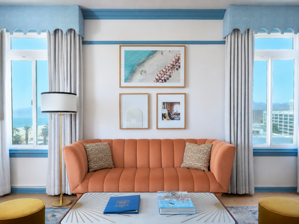 A bright living room with an orange sofa, framed artwork, sheer curtains, big windows, and a standing lamp beside a glass coffee table.