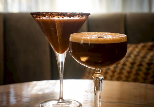 Two amber cocktails in glasses on a bar table, with one in the background and a latte-like foam top in the foreground.
