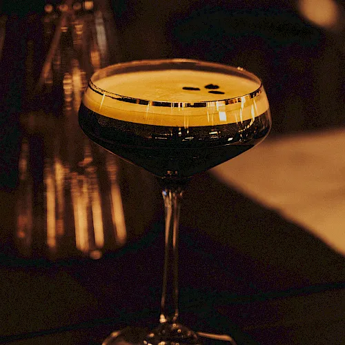A dimly lit bar close-up of a stemmed cocktail glass with an amber drink and bokeh lights in the background.