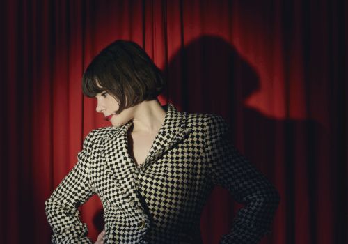 A stylish woman with a dark bob and red lipstick poses in front of red theater curtains, wearing a black-and-cream patterned dress.