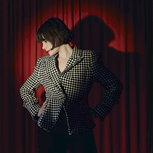 A stylish woman with a dark bob and red lipstick poses in front of red theater curtains, wearing a black-and-cream patterned dress.