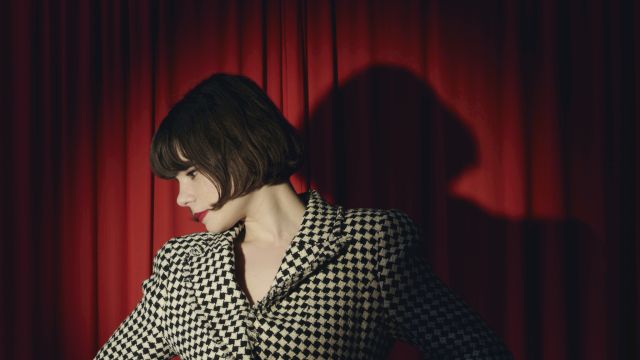 A stylish woman with a dark bob and red lipstick poses in front of red theater curtains, wearing a black-and-cream patterned dress.