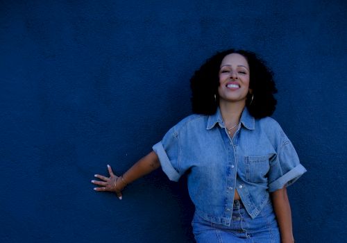 Smiling woman in a denim outfit poses beside a vivid blue wall, flashing a confident grin.