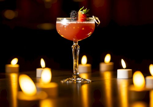 A stemmed cocktail with strawberry and blackberry garnish sits amid warm candlelight, in a dark, cozy bar scene.