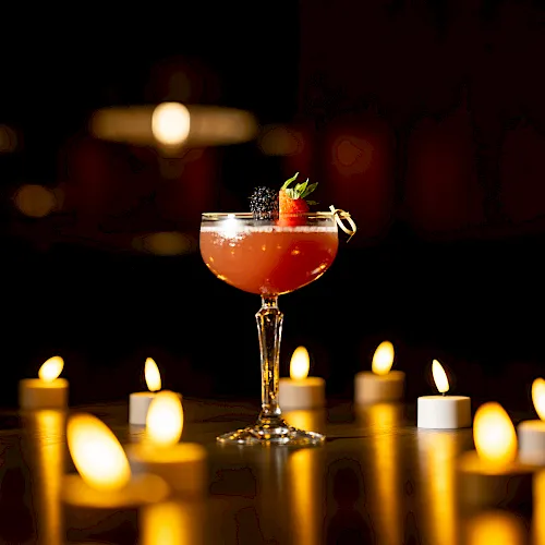 A stemmed cocktail with strawberry and blackberry garnish sits amid warm candlelight, in a dark, cozy bar scene.