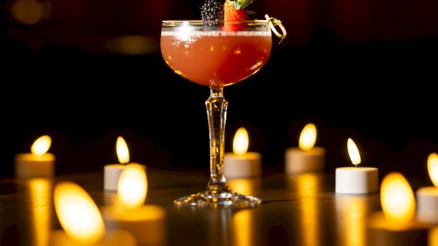 A stemmed cocktail with strawberry and blackberry garnish sits amid warm candlelight, in a dark, cozy bar scene.