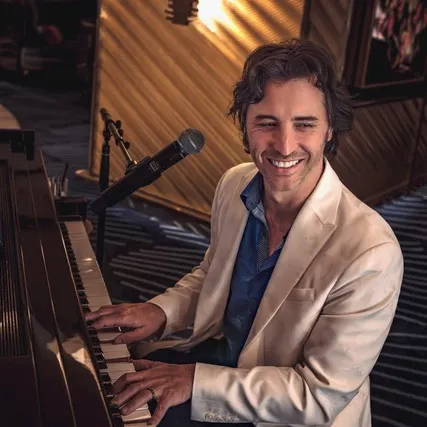 A smiling man in a white blazer plays a grand piano in a studio with microphones and warm lighting.