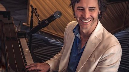 A smiling man in a white blazer plays a grand piano in a studio with microphones and warm lighting.