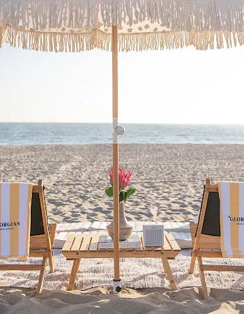 Two striped beach chairs and a fringe umbrella set up on sandy shores with a small flower vase and ocean view.