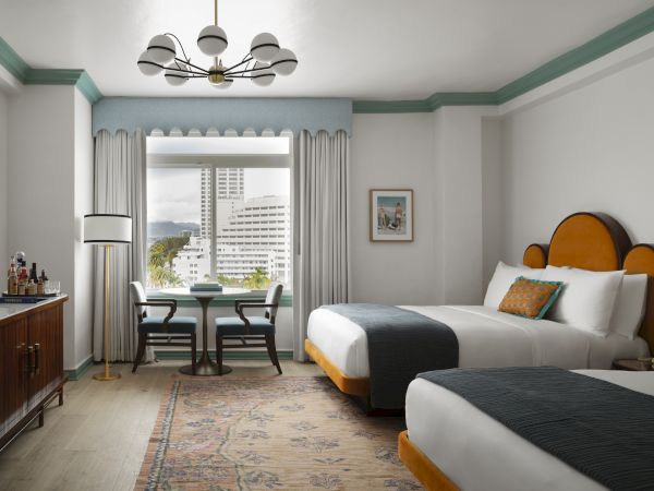 The image shows a hotel room with two beds, a window with a view, a table with chairs, a chandelier, and a bar cart.