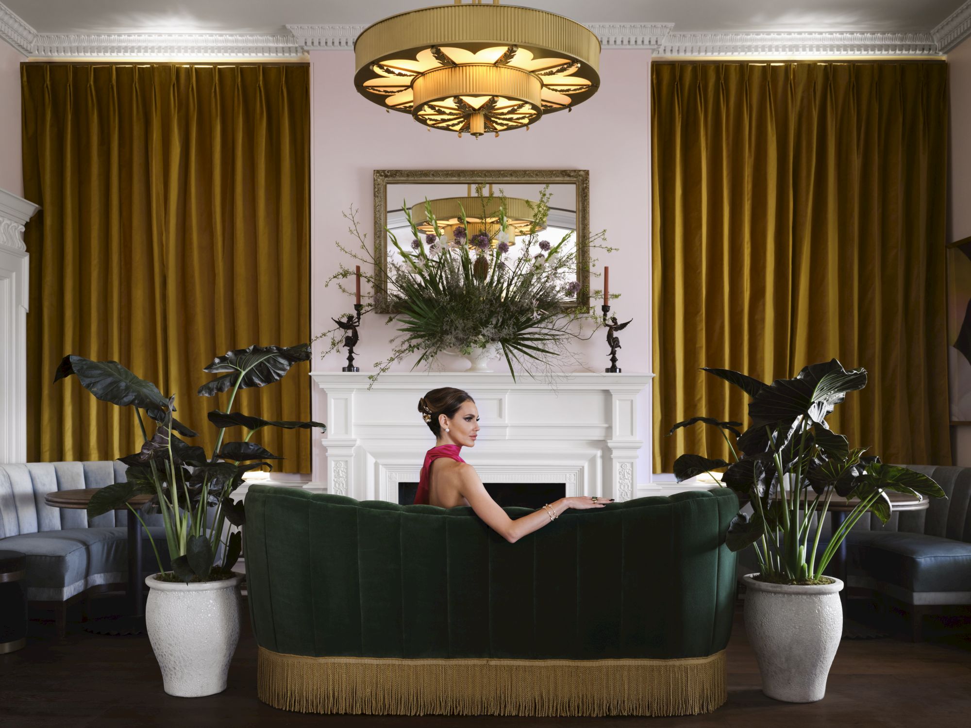 A woman in a red dress sits on a green sofa in an elegant room with yellow curtains, a chandelier, and potted plants.