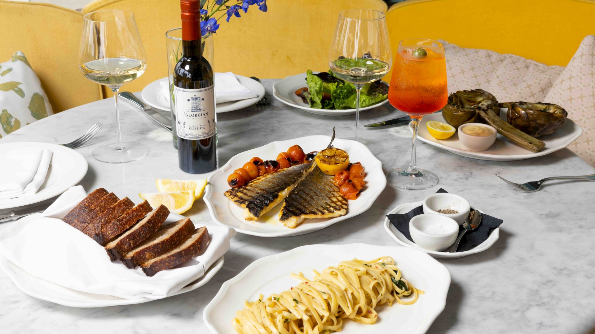 A dinner table set with grilled fish, pasta, bread, salads, two wine glasses, and an orange drink, beside a bottle of wine.
