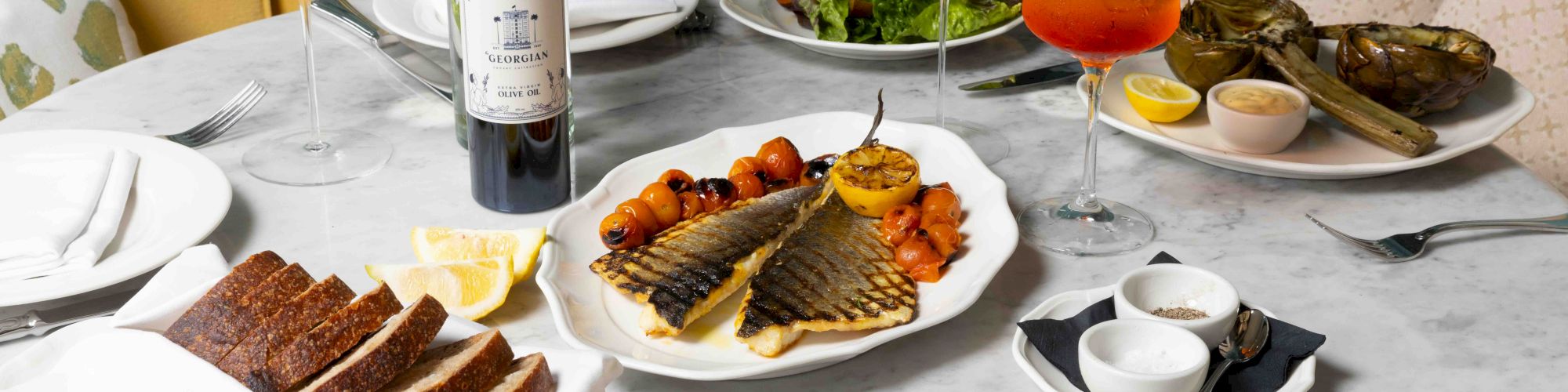 A dinner table set with grilled fish, pasta, bread, salads, two wine glasses, and an orange drink, beside a bottle of wine.