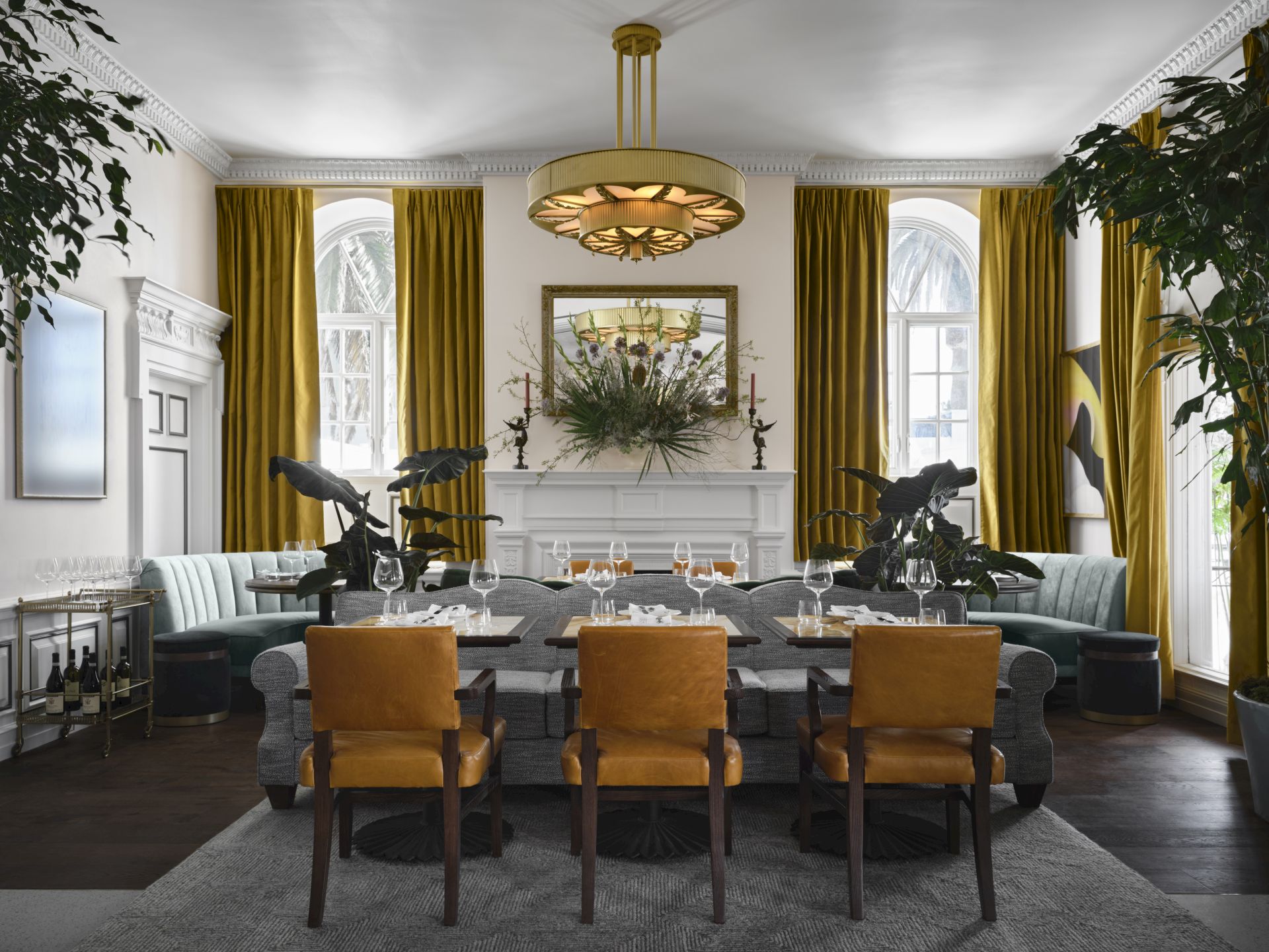 An elegant dining room setup with a central table, gold chandelier, velvet drapes, and floral arrangements amid greenery.