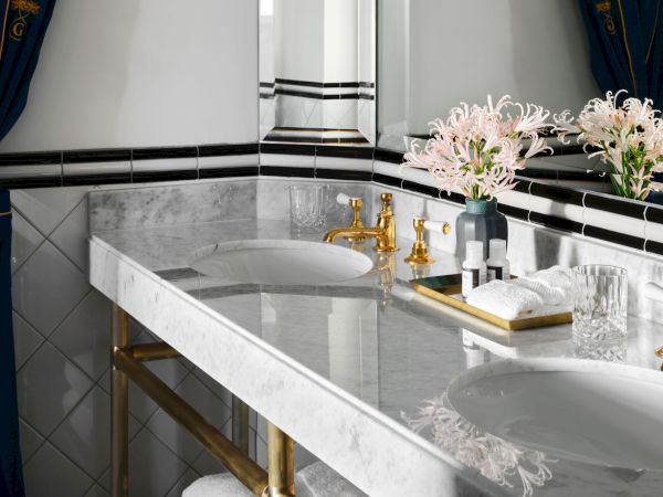 Luxurious bathroom with a marble countertop, twin sinks, gold fixtures, flowers, and rolled towels on a tray. Decorative navy curtains.