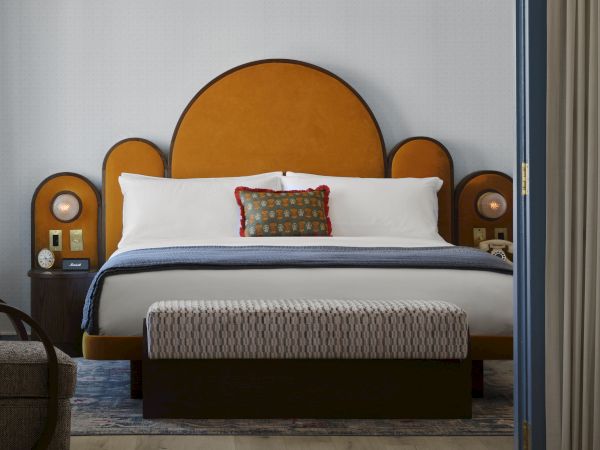The image shows a modern bedroom with an orange headboard, double bed, patterned ottoman, and bedside tables, creating a cozy ambiance.