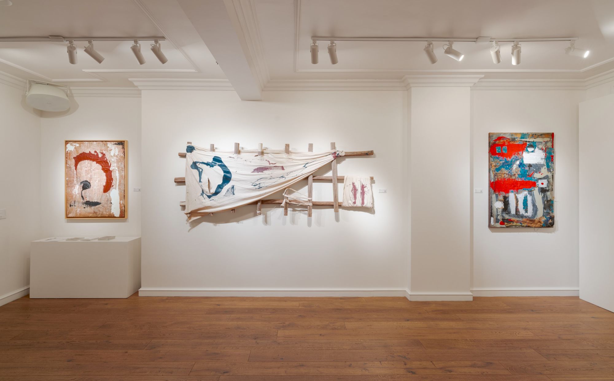 A bright contemporary gallery with white walls and wooden floors, showcasing three abstract artworks, one with a red circle, a central wooden sculpture, and a blue-red piece on the right.