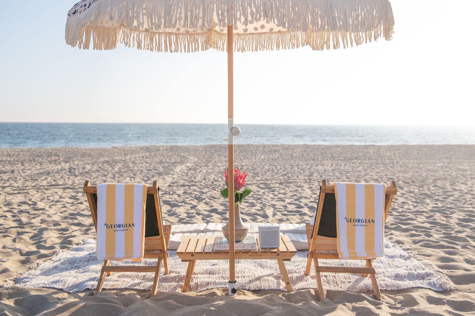 Two striped beach chairs under a fringed umbrella face the sea, with a small table, plant, and book on a sandy beach.