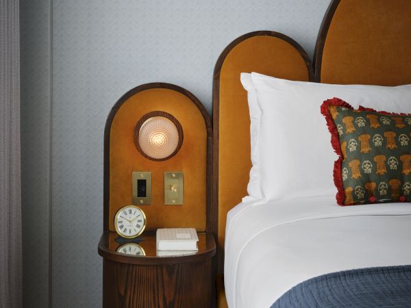A stylish bedside setup with a retro clock, books, and a lamp. Cozy bedding features a decorative pillow with a red fringe edge.