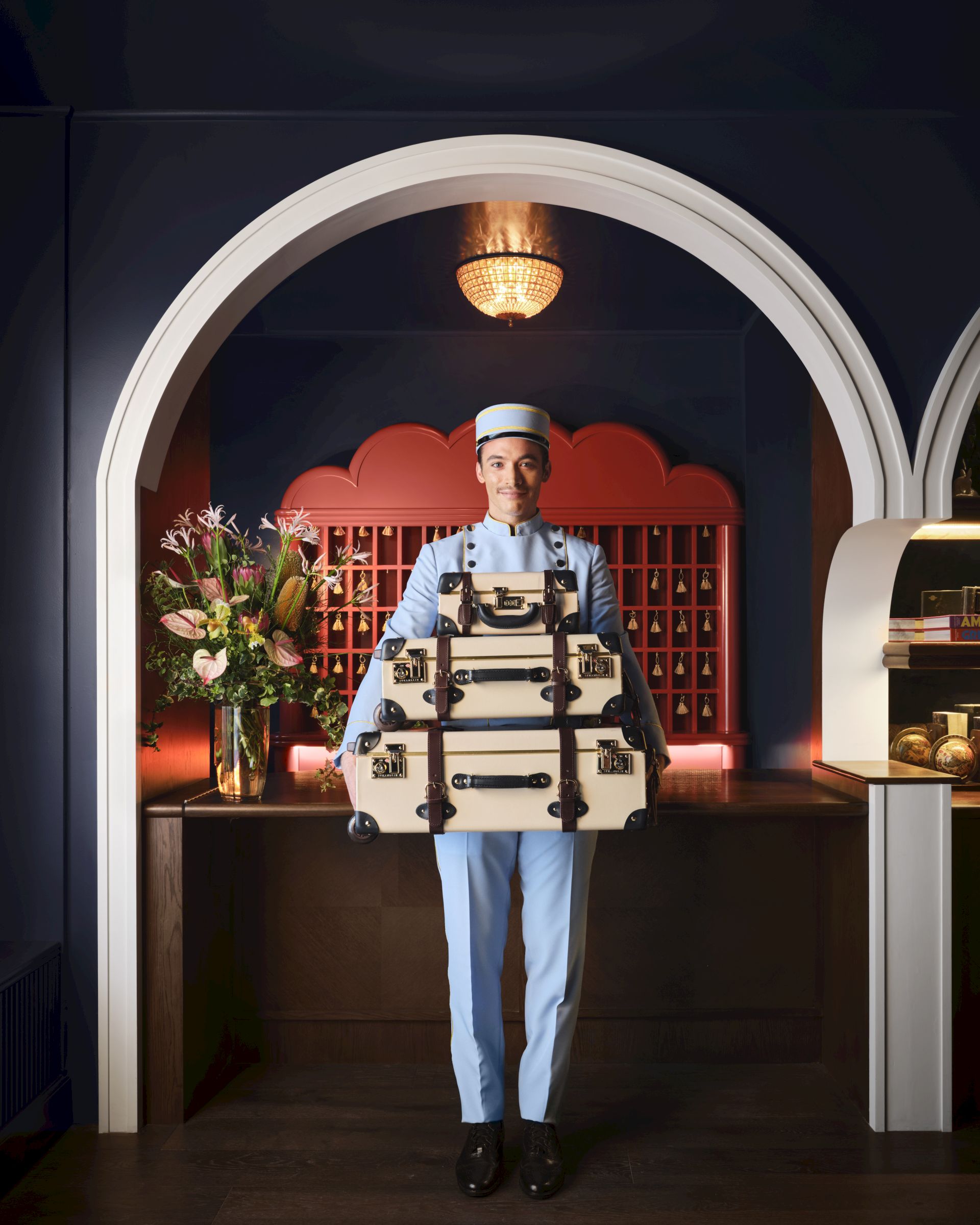 A person in a uniform holds multiple suitcases, standing in an arched doorway with flowers and shelves in the background.