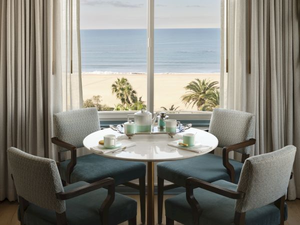 A round table with cups and plates is surrounded by chairs, near a large window with an ocean view and palm trees in the background.