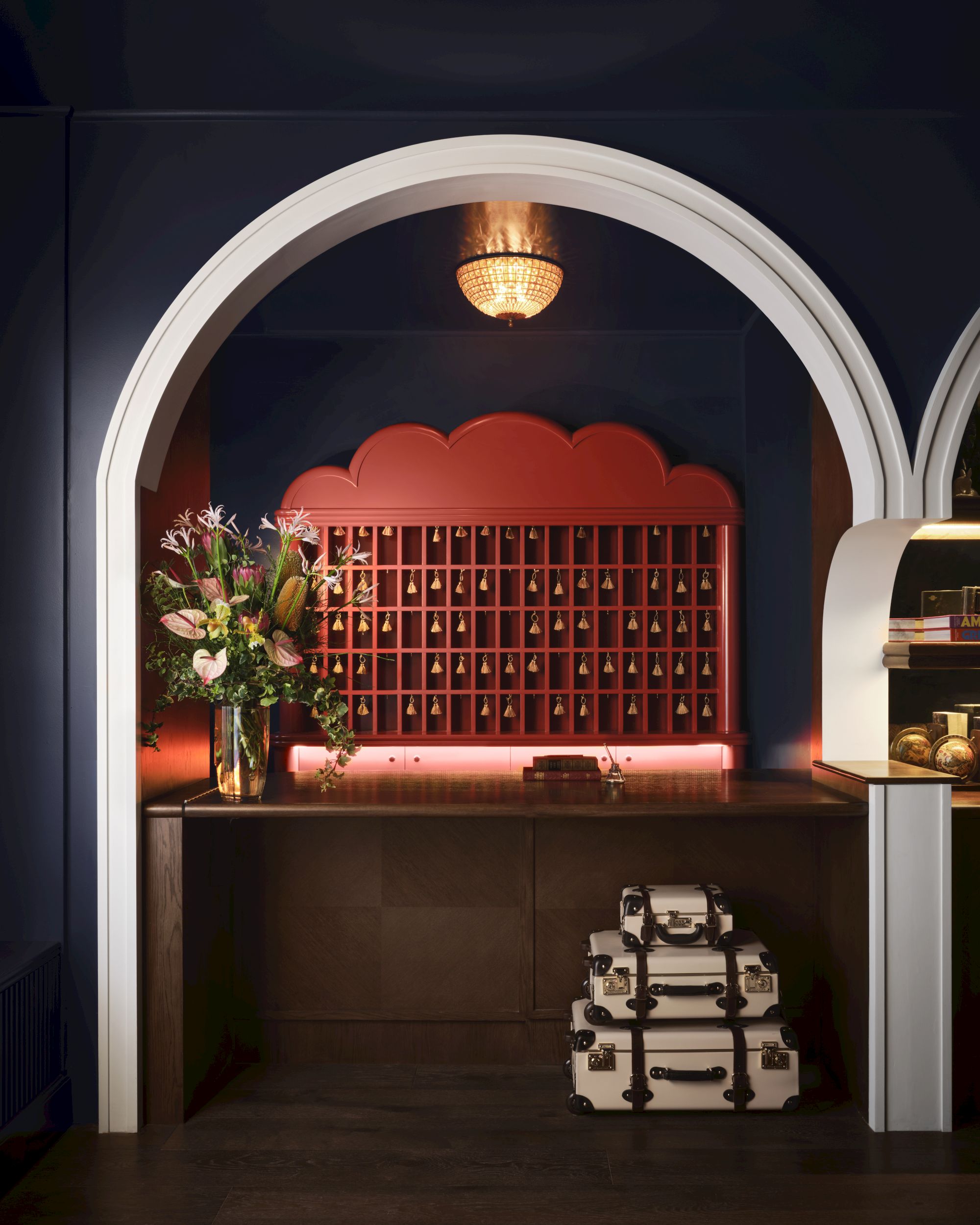 An arched alcove with a reception desk, vintage suitcases, a flower arrangement, and decorative shelving in a dimly lit setting.