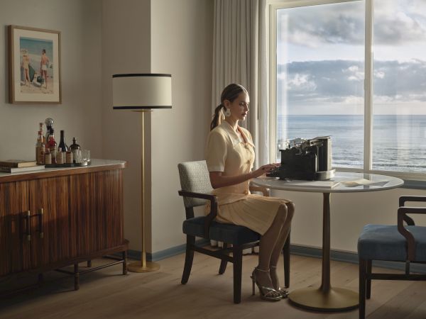 A woman sits at a table by a window with an ocean view, typing on a typewriter. A sideboard and floor lamp are also visible.