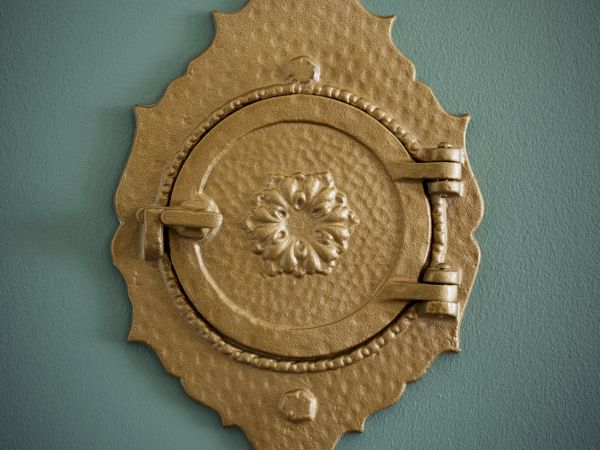 The image shows a decorative, round brass peephole or door viewer with an ornate design on a textured, greenish wall.