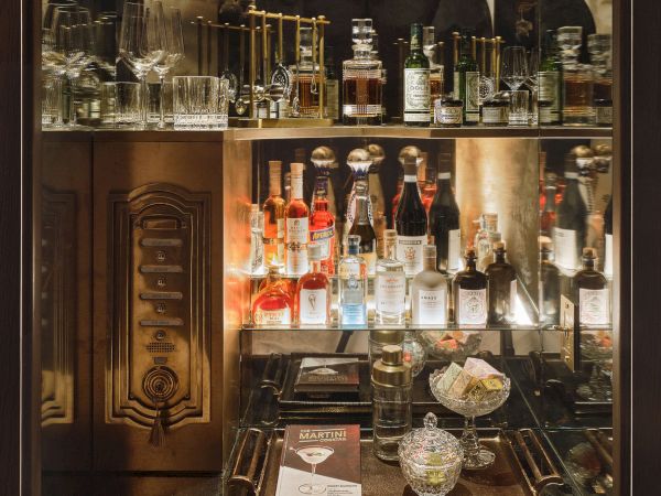 The image shows a well-stocked bar shelf with various bottles of liquor, glasses, and decanters, alongside decorative items and a menu.