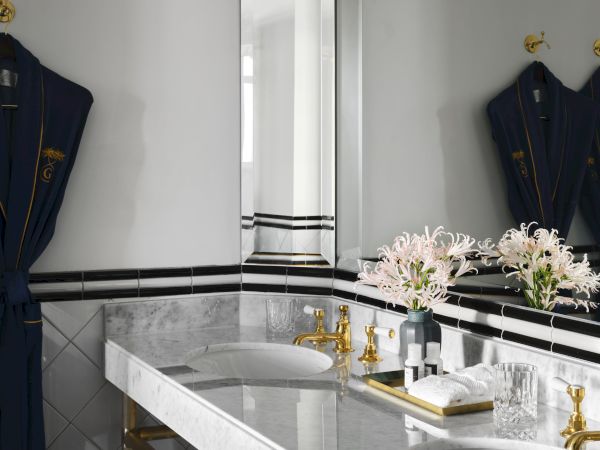 A luxurious bathroom with marble countertops, dual sinks, gold fixtures, a mirror, and flowers on the counter, with robes hanging on hooks.