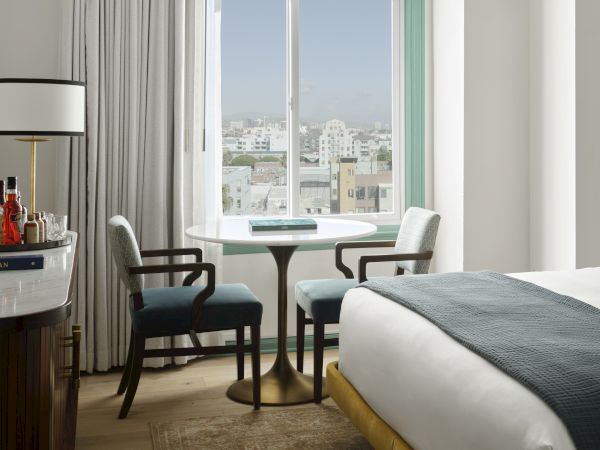 The image shows a cozy hotel room with a bed, a round table, two chairs, a window view, and a countertop with bottles and a lamp.