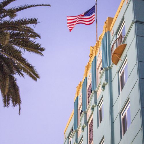 A palm tree and a colorful building with windows are seen beneath a U.S. flag on a pole against a clear sky.