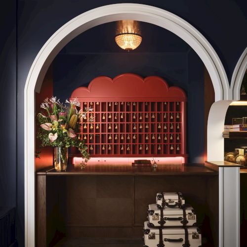 The image shows a stylish hotel reception area with an archway, floral arrangement, red mail slots, and stacked vintage suitcases.