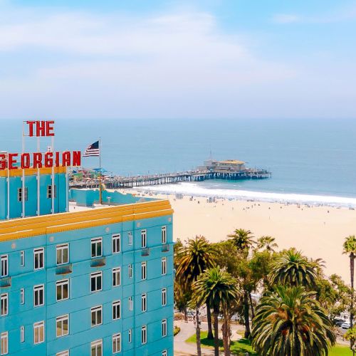 The image shows a seaside view with The Georgian building, palm trees, and a pier extending into a blue ocean under a partly cloudy sky.